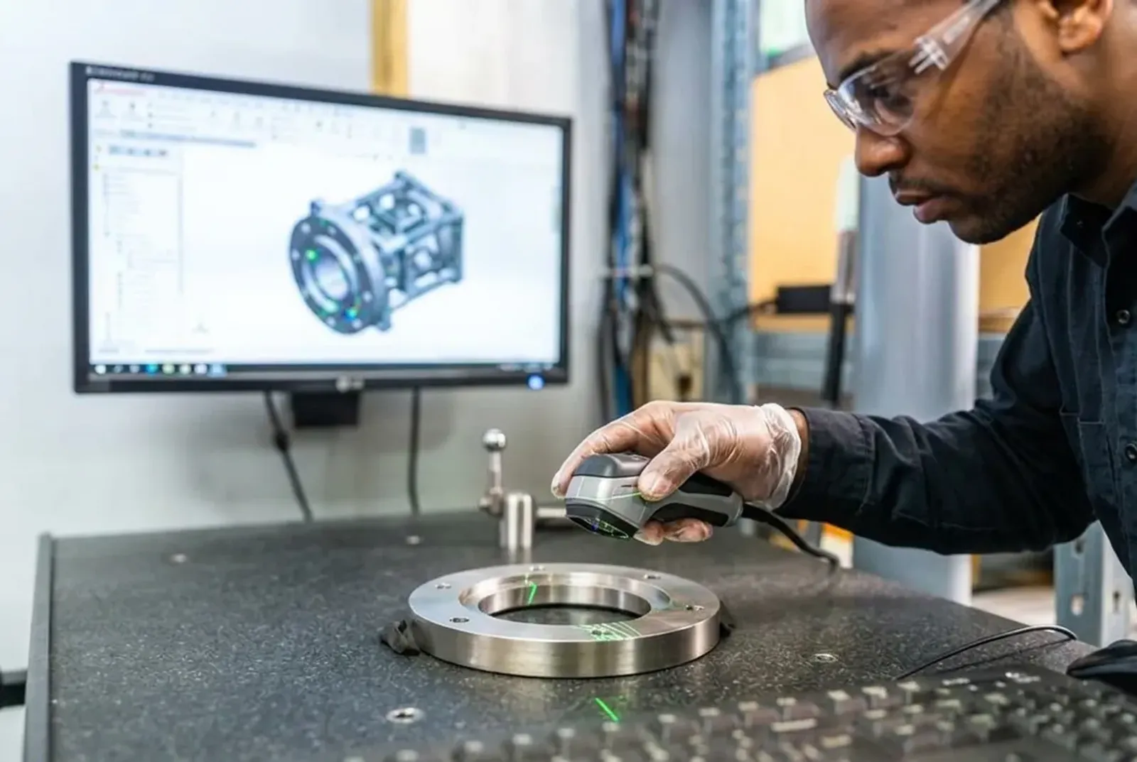 Hand positioning a 3D scanner over a metal part with a CAD model on screen